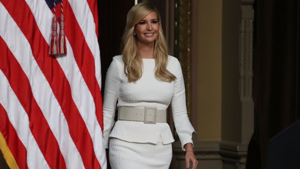 Ivanka Trump smiling in a white outfit while walking by the American flag