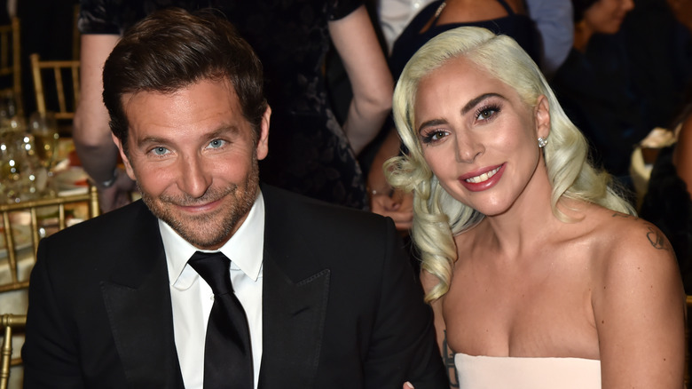 Bradley Cooper in a black suit and tie seated with Lady Gaga in a white strapless gown