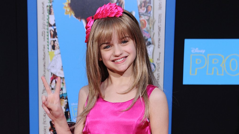 Joey King giving a peace sign and smiling on the red carpet, wearing a pink dress and headband