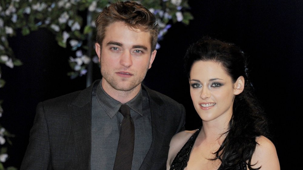 Robert Pattinson with a neutral expression in a dark grey suit, Kristen Stewart smiling in a black dress