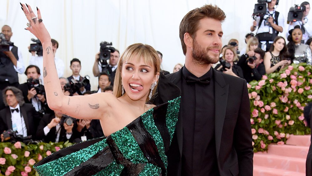 Miley Cyrus in a green-and-black dress, smiling with her tongue out as Liam Hemsworth, in an all-black suit, looks the other way