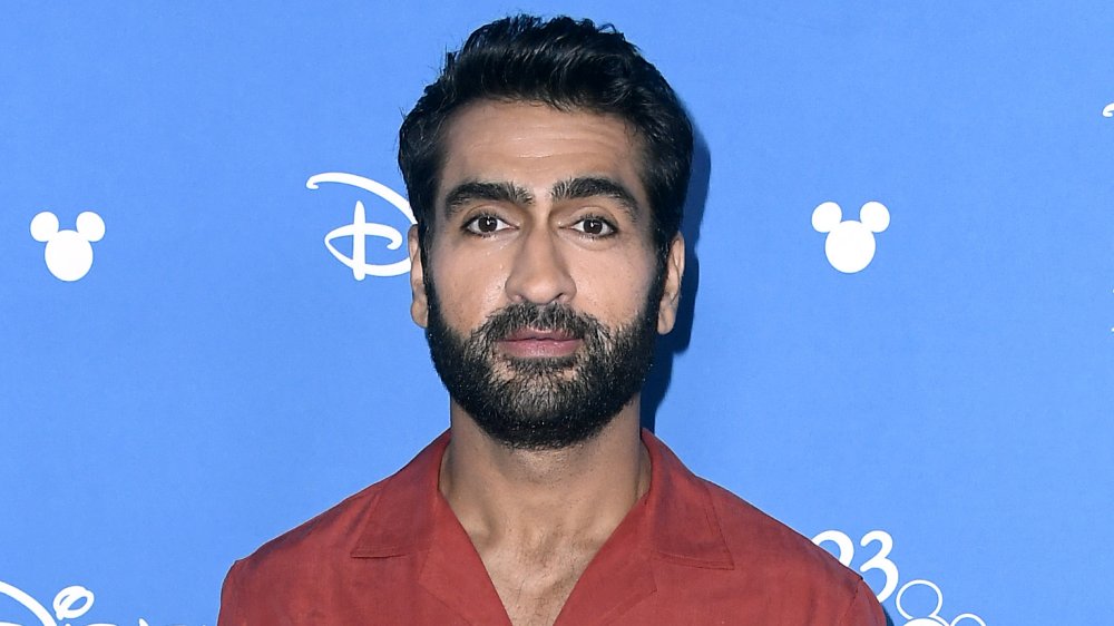 Kumail Nanjiani in a red button-up, posing at a Disney event with a neutral expression
