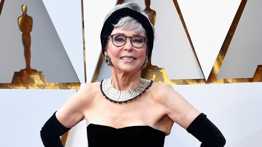 Rita Moreno in a black dress, smiling and posing with both hands on her hips at the Oscars