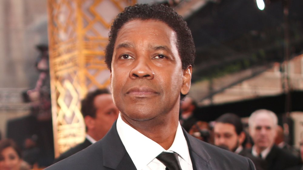 Denzel Washington in a classic black suit with a white shirt and long black tie, posing with a neutral expression