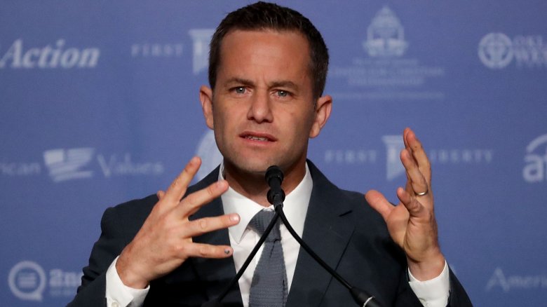 Kirk Cameron in a grey suit, looking concerned while speaking with his hands at a podium