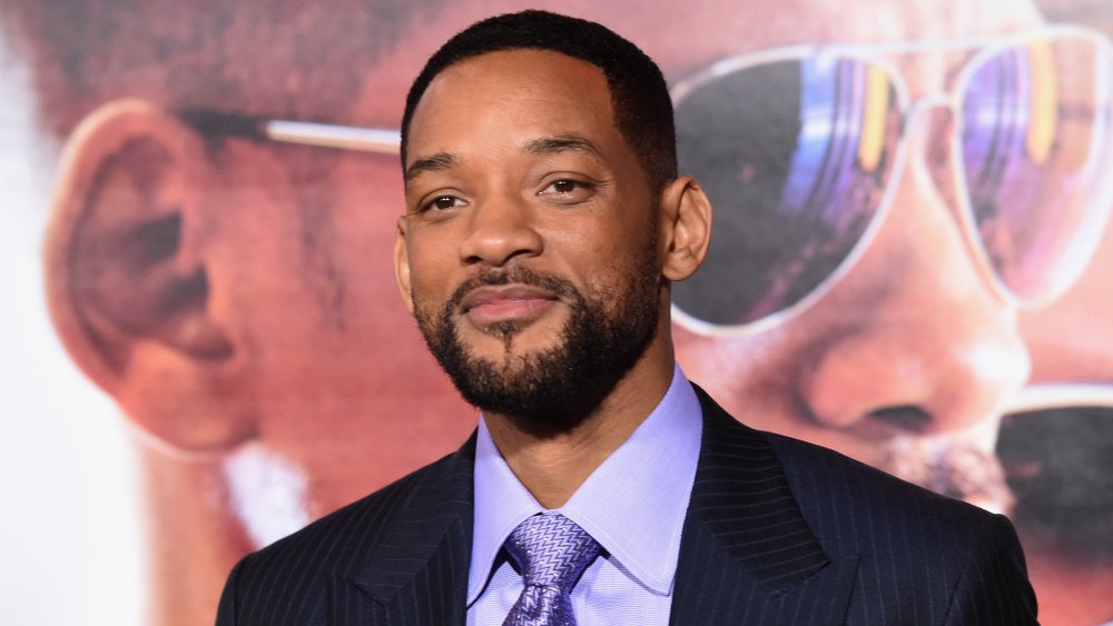 Will Smith in a black pin-striped suit and purple tie, posing with a small smile at a movie premiere