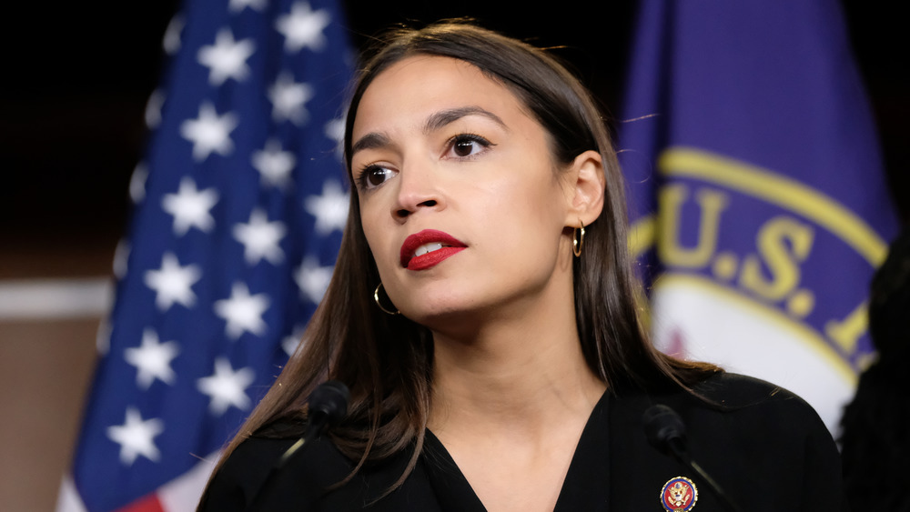 Alexandria Ocasio-Cortez speaking with a serious expression in front of the American flag