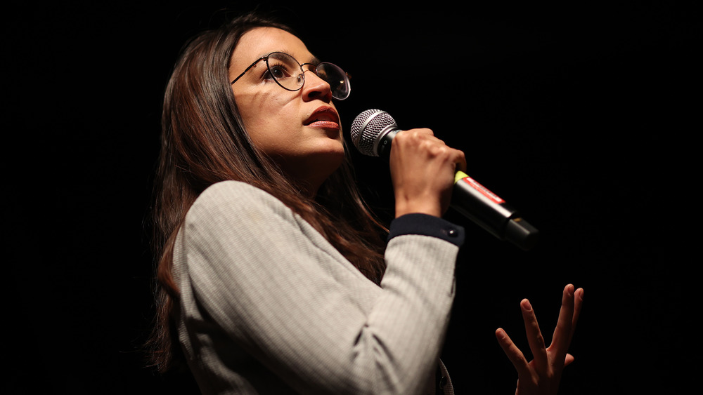 Alexandria Ocasio-Cortez speaking into a microphone with a serious expression