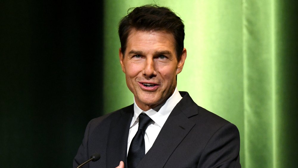 Tom Cruise speaking at a podium, in front of green wall