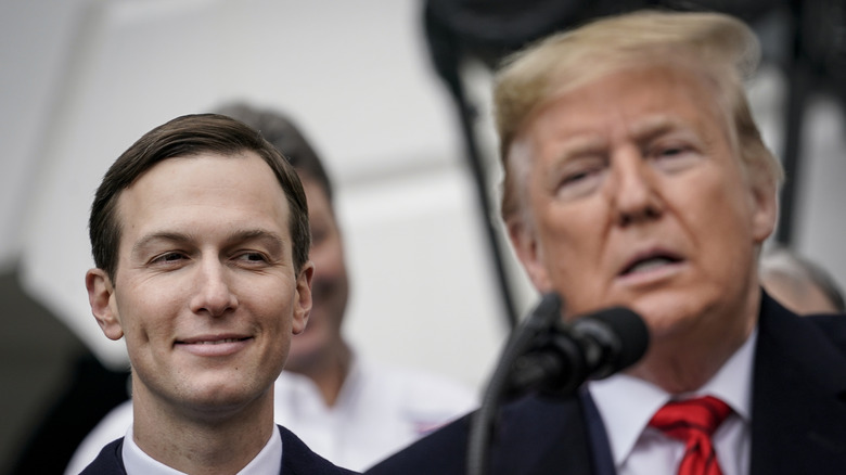 Jared Kushner smiling while standing behind Donald Trump