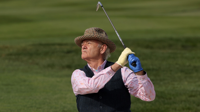 Bill Murray wear a pink top and vest as he swings his club on the golf course