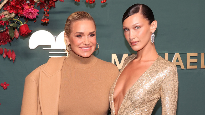 Yolanda Hadid wearing a tan turtleneck and jacket with Bella Hadid wearing a gold low cut sparkling gown