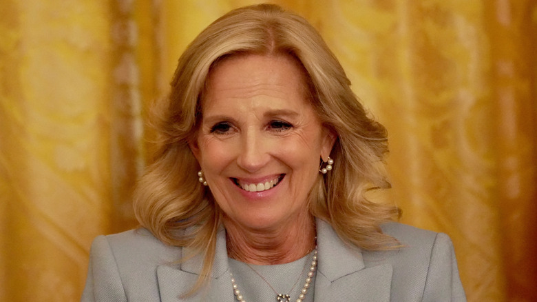 Jill Biden smiling in a gray outfit with pearl earrings