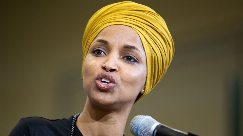 Ilhan Omar wearing a yellow hijab and black outfit, speaking passionately into a mic