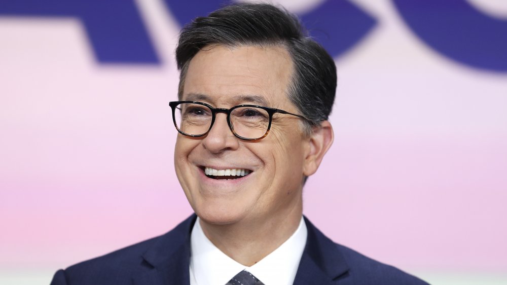 Stephen Colbert in a navy blue suit, smiling big while looking off to the side