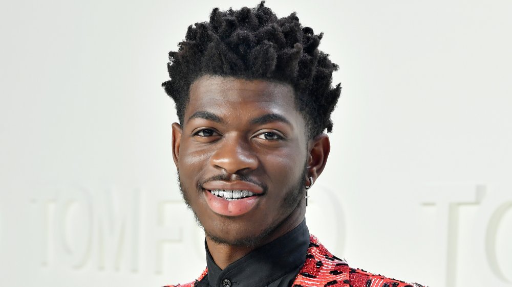 Lil Nas X wearing a diamond cross earring, teeth grills, and a red-and-black suit, smiling