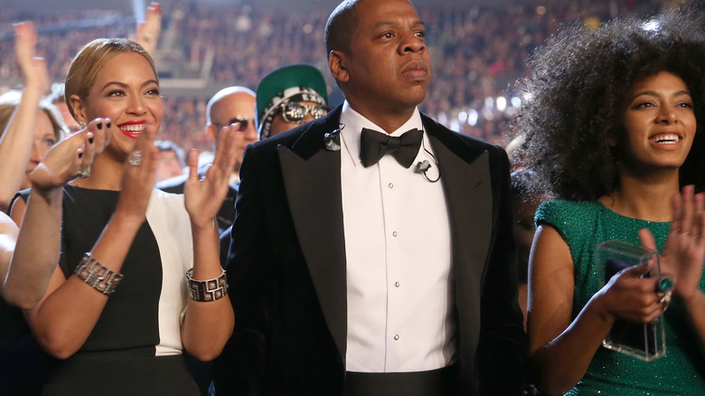 Jay-Z looking worried next to clapping Beyonce and Solange Knowles