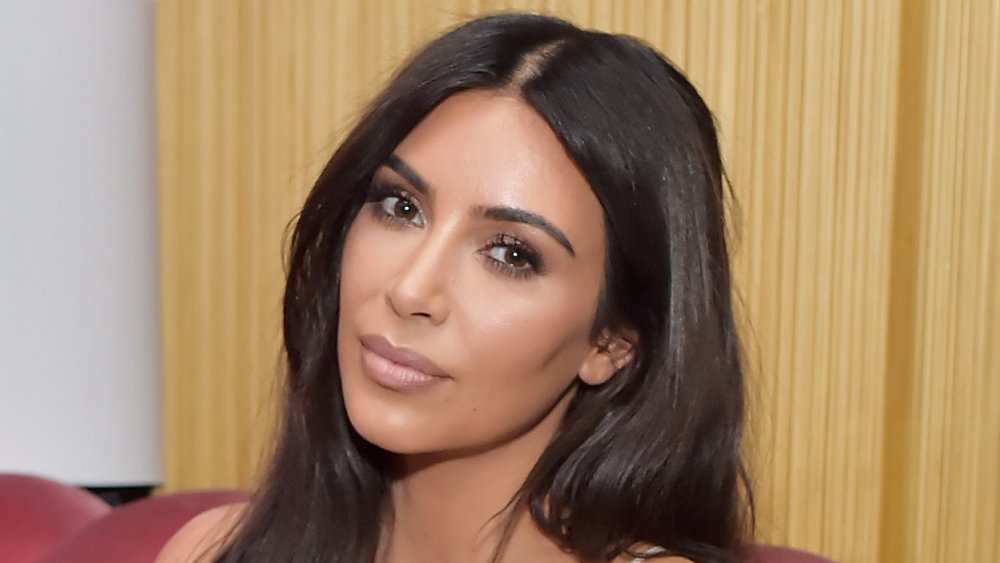 Kim Kardashian in a white dress, posing on a couch with her head tilted with a neutral expression