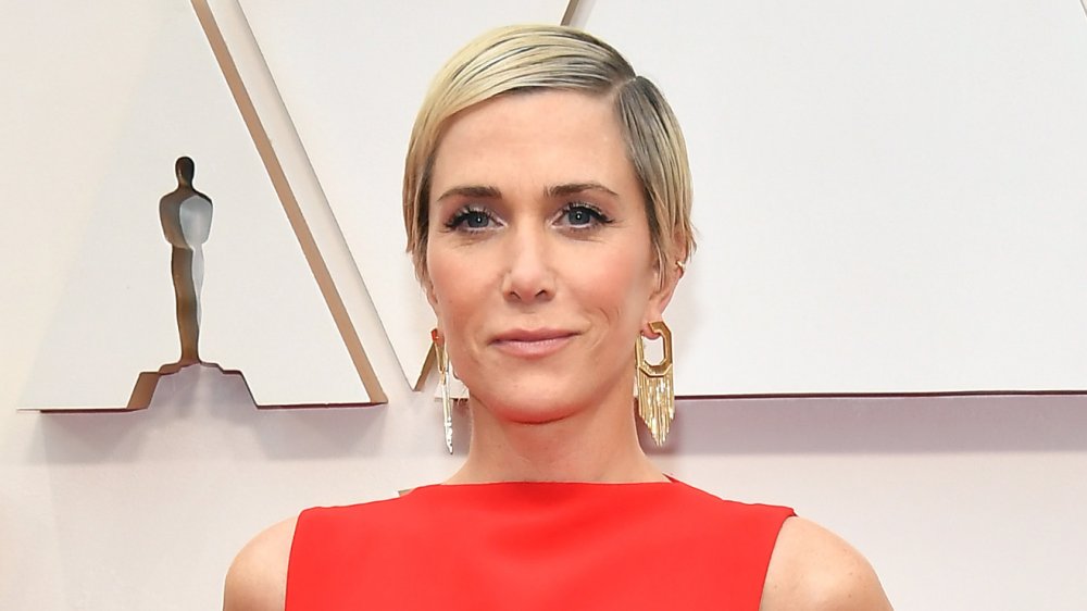 Kristen Wiig in a red dress, smirking on the Oscars red carpet