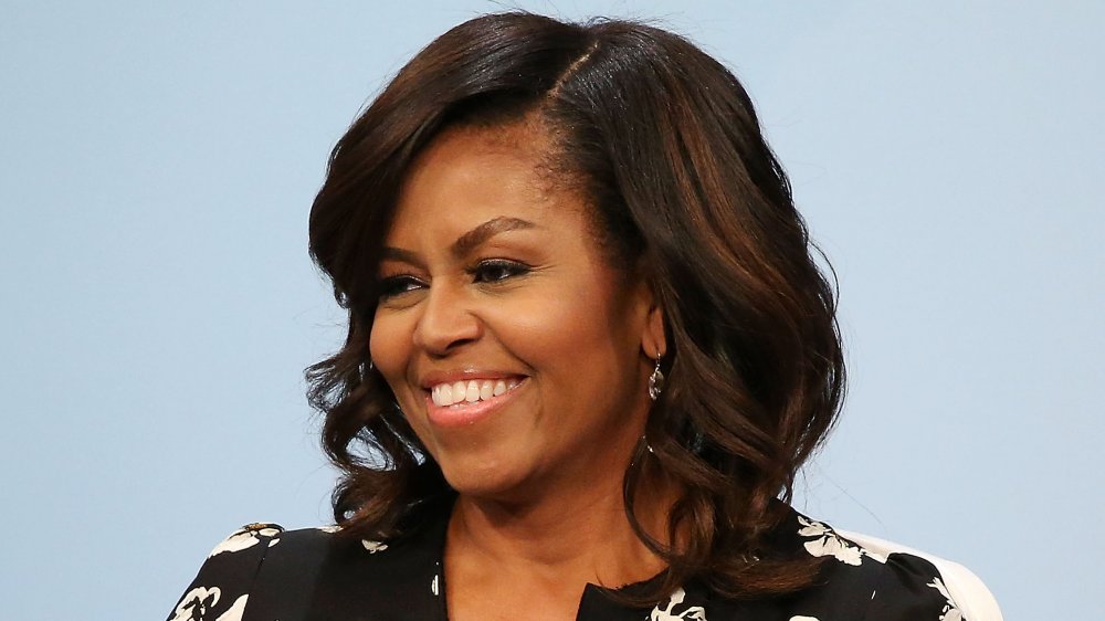 Michelle Obama in a black floral-printed dress, smiling while looking off to the side