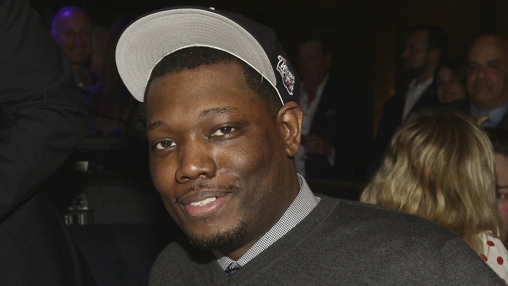Michael Che in a gray sweater and black hat, posing with a small smile