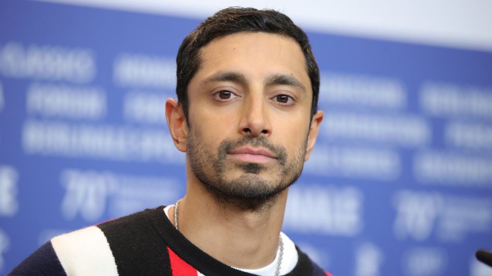 Riz Ahmed in a black, red, and white sweater, posing during an interview with a neutral expression