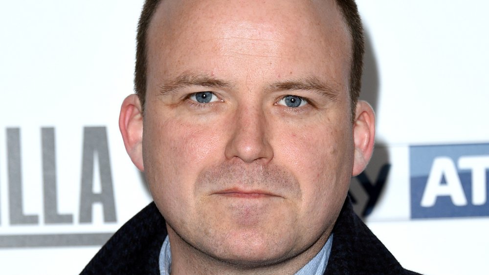 Rory Kinnear in a black-and-blue checkered coat, posing with a neutral expression