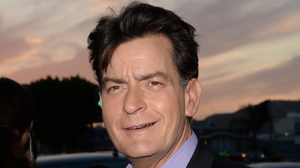 Charlie Sheen with a small smile, wearing a black blazer and blue shirt against a sunset