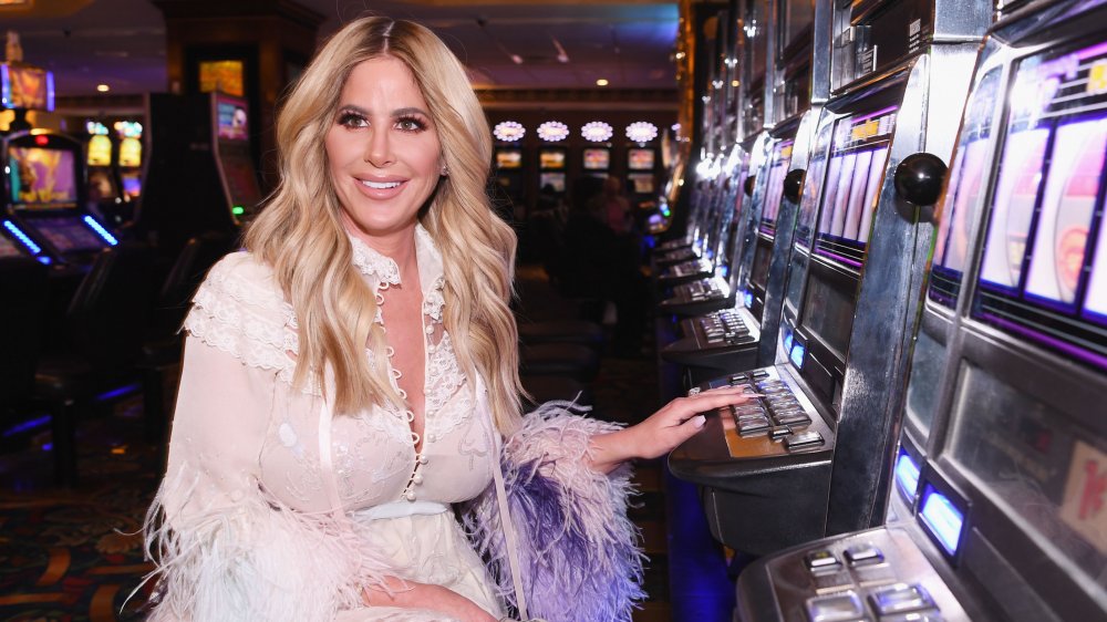 Kim Zolciak-Biermann in an all-white, lacy outfit, smiling while sitting at a casino slot machine