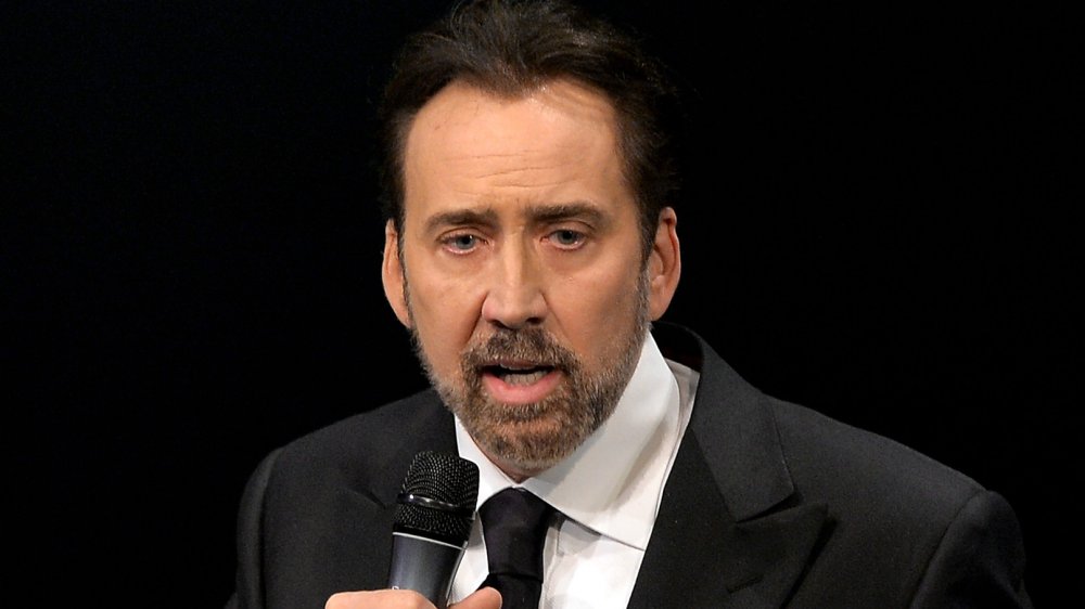 Nicolas Cage wearing a classic black suit and tie, sporting a beard, speaking into a mic