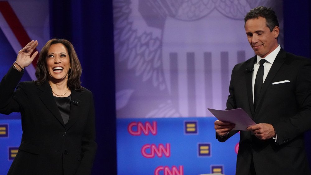 Kamala Harris waving on stage and Chris Cuomo looking at his notes next to her