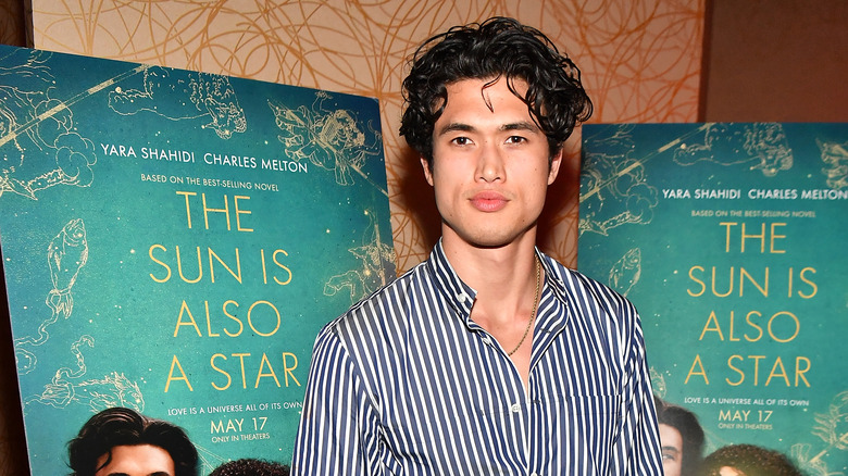 Charles Melton posing in front of two film posters for The Sun Is Also a Star