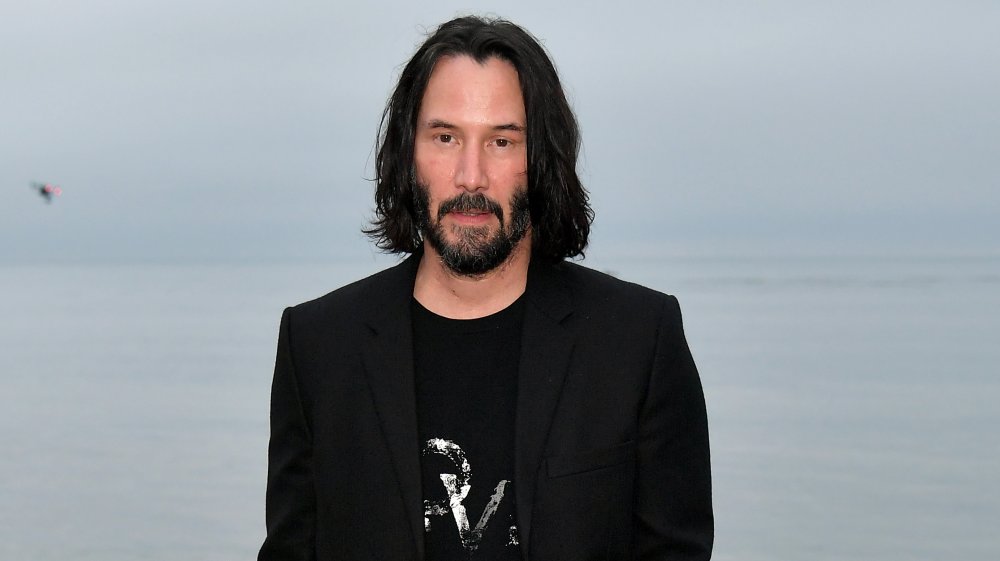 Keanu Reeves in a long, black blazer and black t-shirt, with shoulder-length hair, posing on the beach