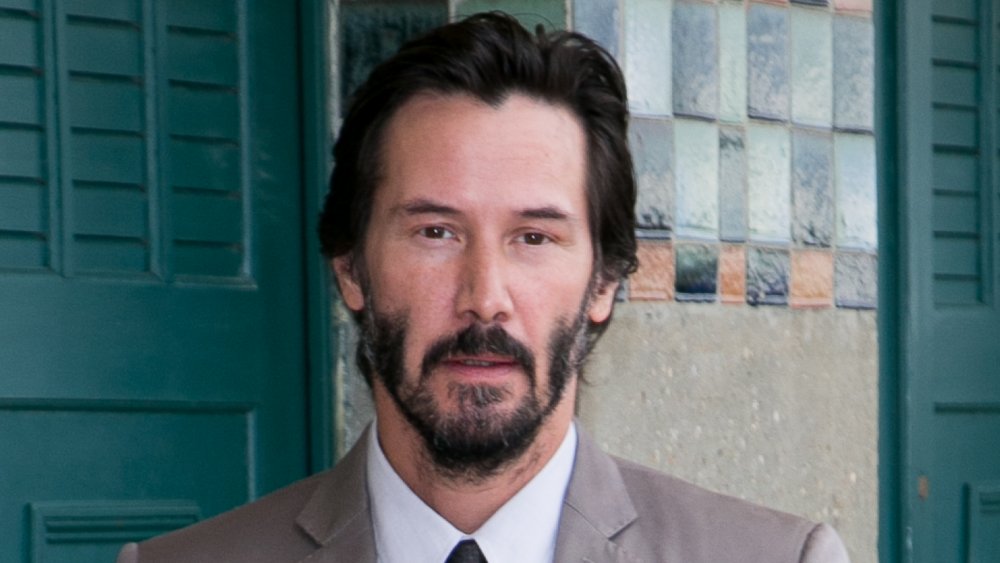 Keanu Reeves in a tan suit and white shirt, with shorter hair, looking straight at the camera with a neutral expression