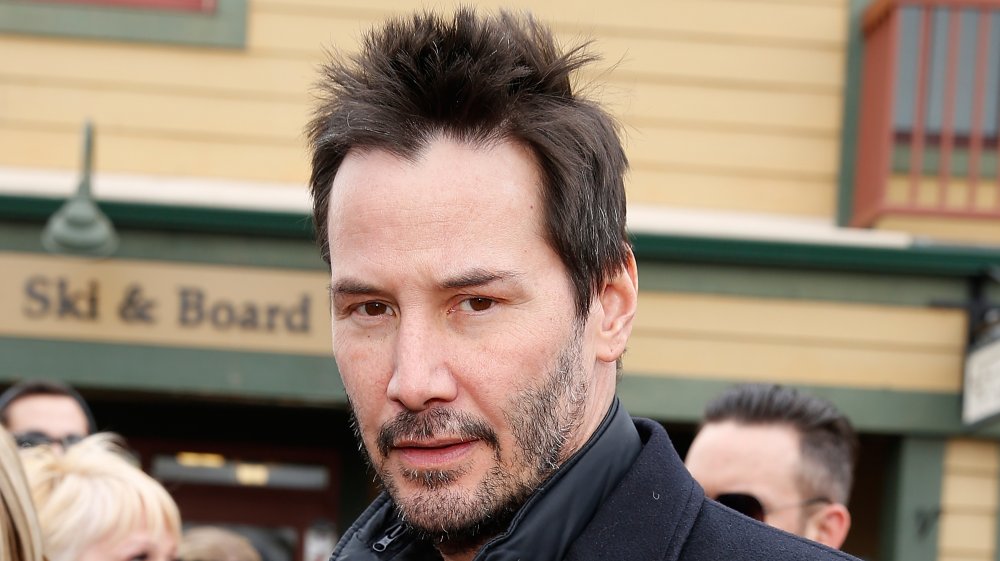 Keanu Reeves wearing a black pea coat, with shorter, spiky hair, looking straight at the camera with a serious expression