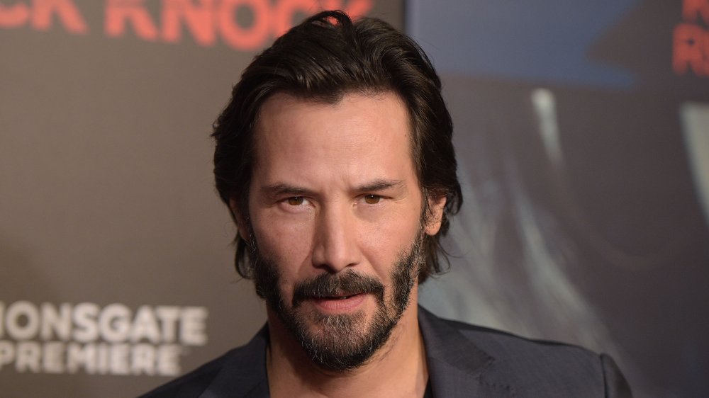 Keanu Reeves in a dark grey blazer and black shirt, with shorter hair, at a premiere