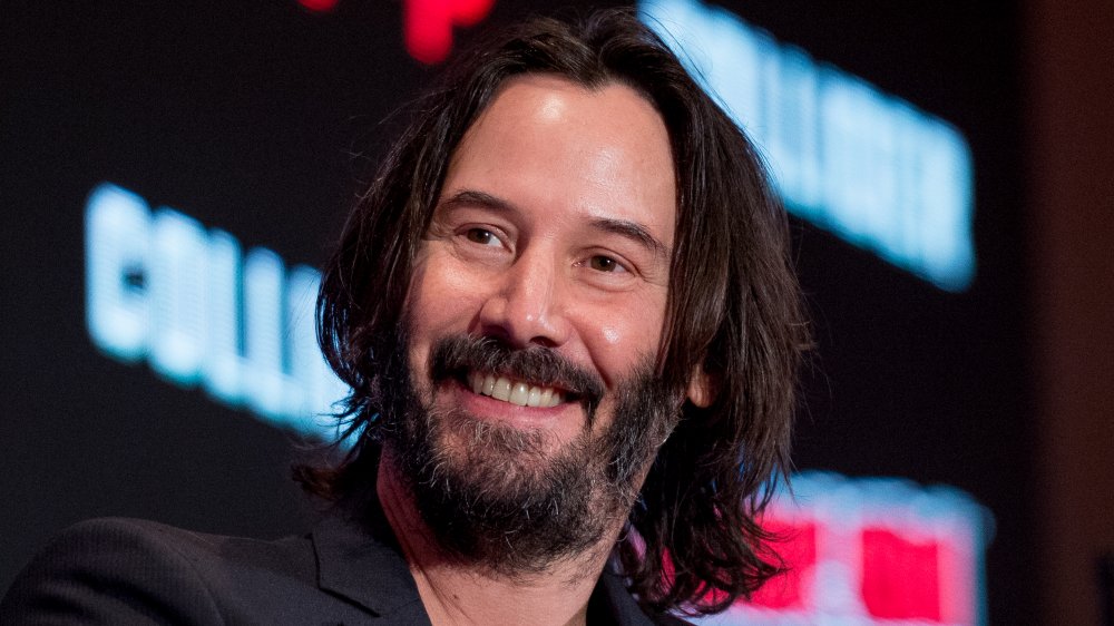 Keanu Reeves wearing a black blazer and v-neck shirt, smiling during a Comic Con panel