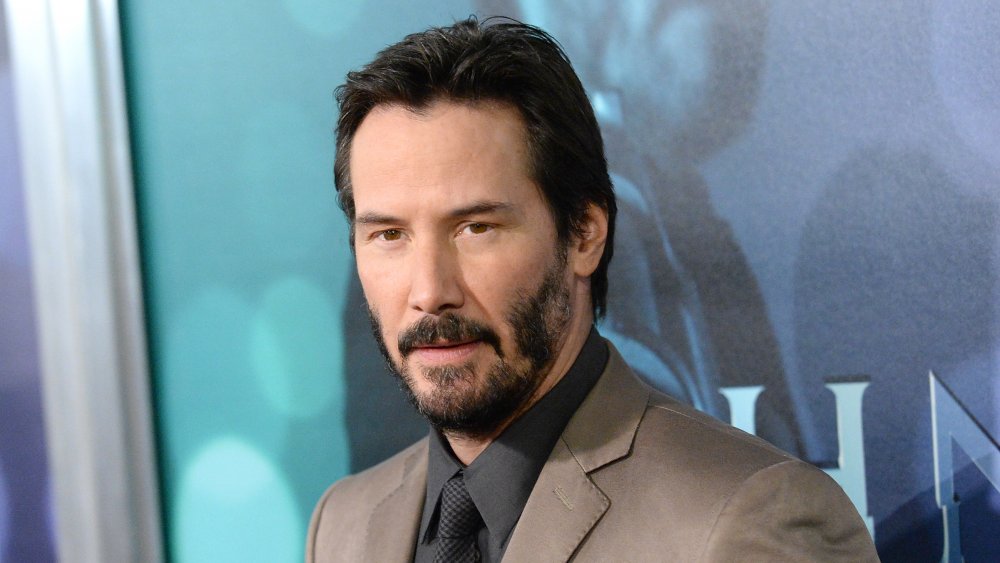 Keanu Reeves wearing a tan suit and black shirt, with short hair, at a movie premiere