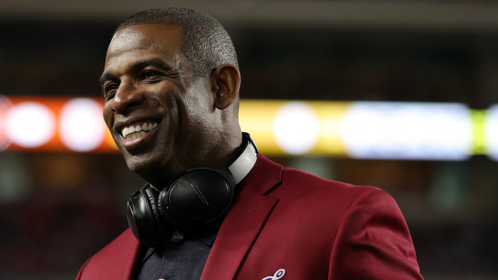 Deion Sanders smiling, while looking off to the side, in a maroon blazer and black polo shirt