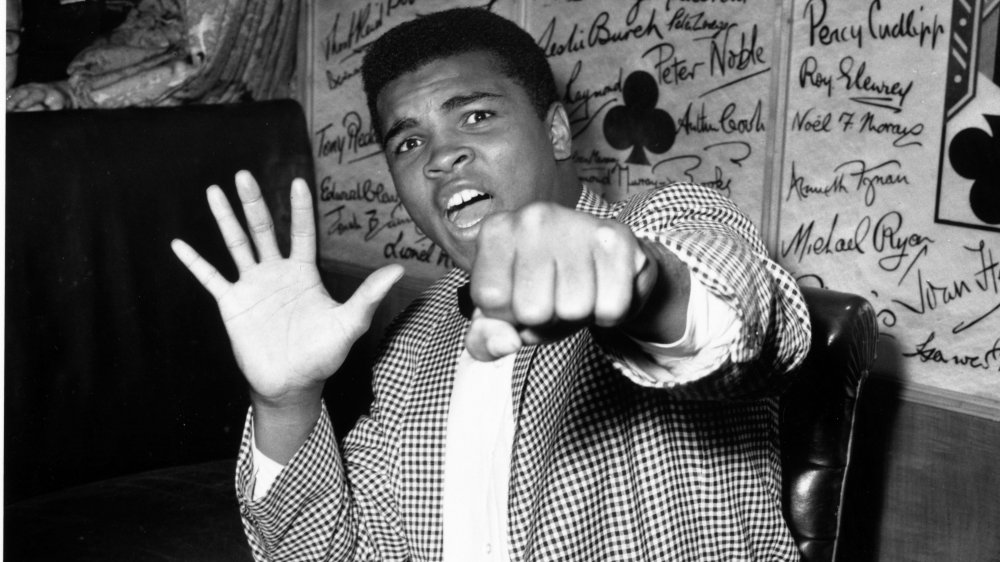 Black-and-white photo of Muhammad Ali in street clothes, striking a playful boxing pose
