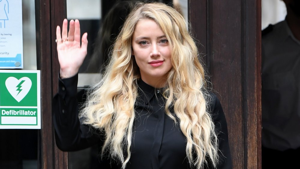 Amber Heard in a black outfit, walking outside with a smirk and waving