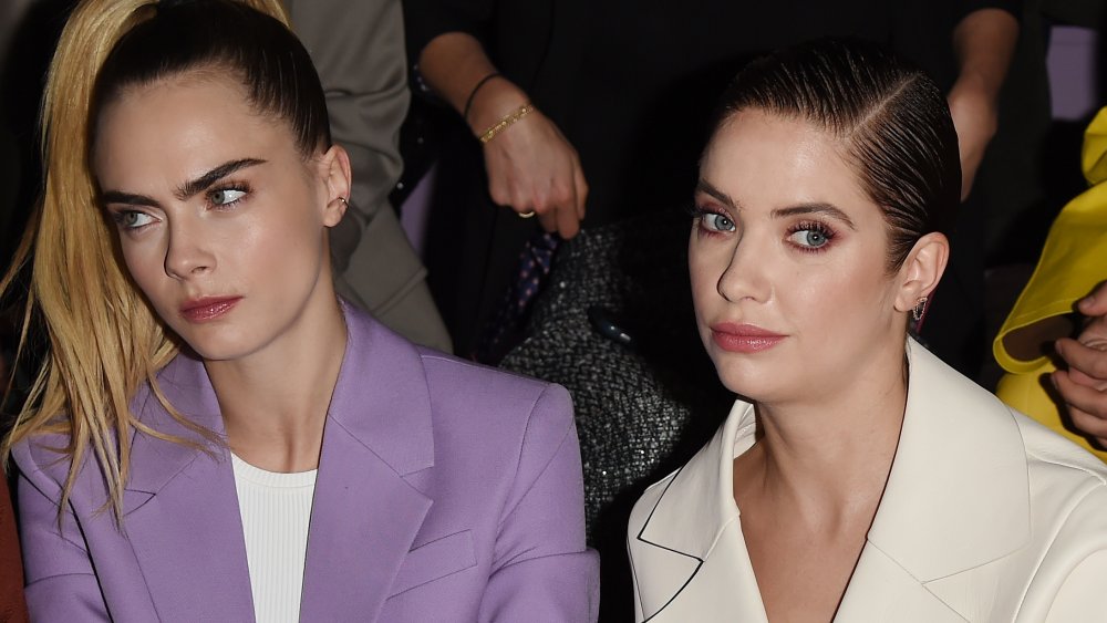 Cara Delevingne in a purple blazer, Ashley Benson in a white blazer, both looking in different directions with unhappy expressions