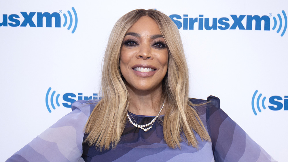 Wendy Williams wears a purple wavy-stripped sweater and pearls while smiling for a photo