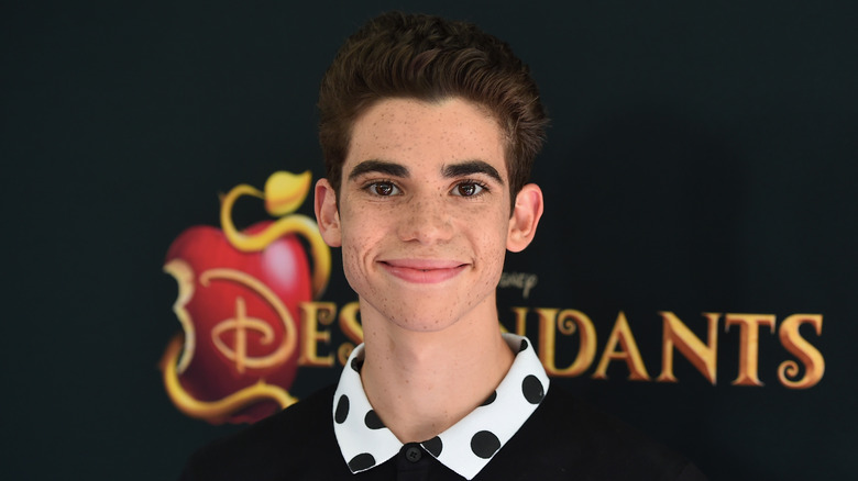 Cameron Boyce grinning in front of Descendants logo