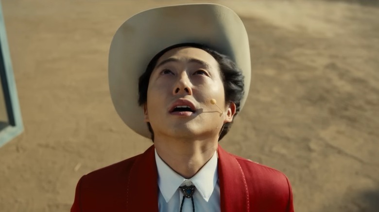 Steven Yeun in a cowboy hat, a red jacket, looking up