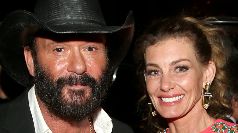 Tim McGraw and Faith Hill pose at an event as McGraw rocks a beard and cowboy hat and Hill wears her hair in a ponytail with yellow chunky earrings on