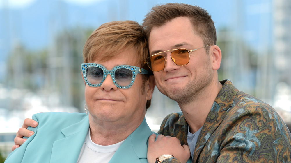 Elton John, at Cannes, smirking, wearing blue sparkly glasses, a blue suit; Taron Egerton, smiling, wearing sunglasses, in a pattern shirt, Cannes 2019