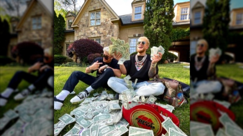 Jake Paul in sunglasses on a front lawn of a house holding dollar bills in his hand next to unidentified companion