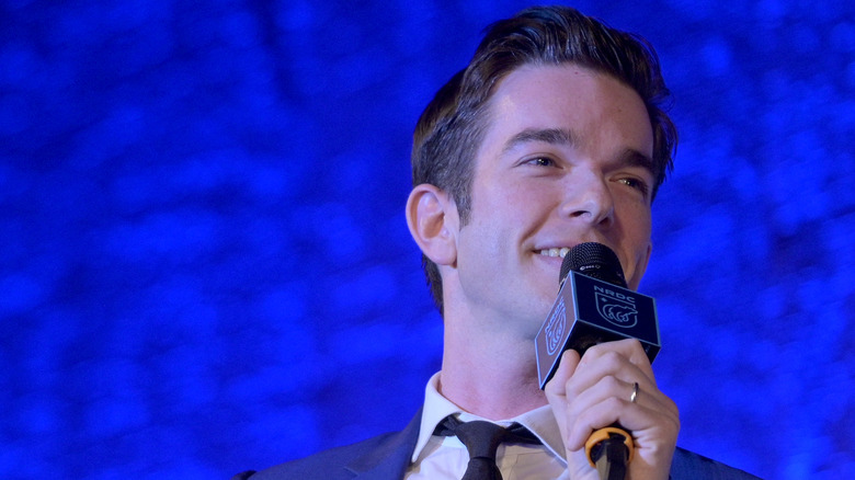 John Mulaney smiling and speaking into a microphone wearing a tie
