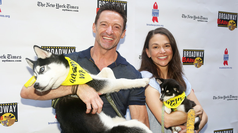 Hugh Jackman and Sutton Foster smiling while holding dogs wearing yellow bandanas